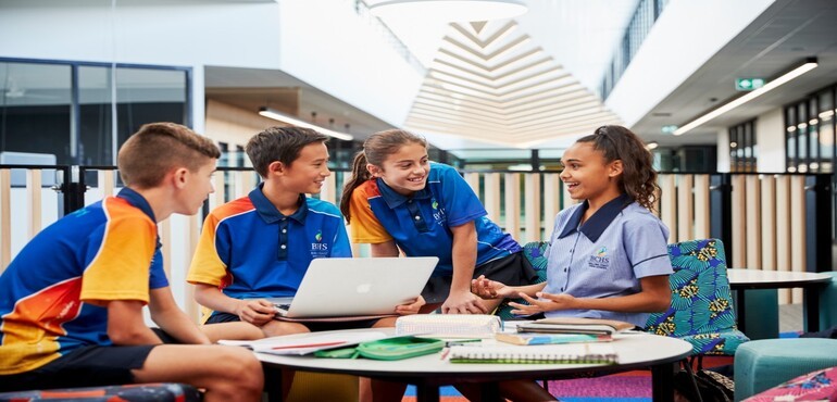 Four high school students talking while sitting at a table with a laptop and notebooks.